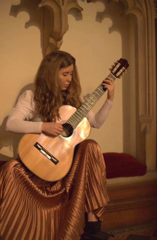 India Falconer playing classical guitar beneath gothic stone arches in candlelight