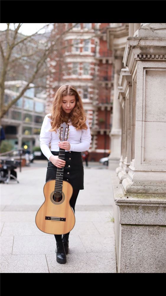 India Falconer standing with her classical guitar beside a stone building