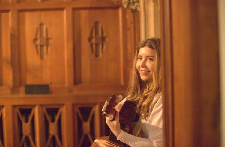India Falconer performing at a church concert, smiling with her guitar among warm wood panelling